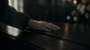 A hand rests on a polished wooden coffin in soft natural light