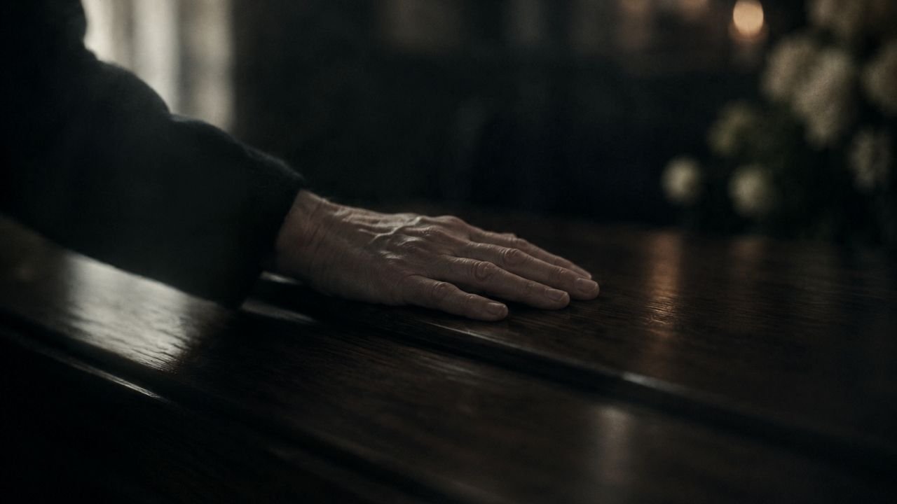 A hand rests on a polished wooden coffin in soft natural light