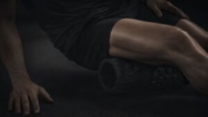 A person uses a black foam roller beneath one thigh, with one hand on the floor