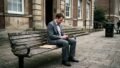 A man in a grey suit sits on a wooden bench, looking at his phone, with a manila folder beside him