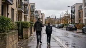 A father and his young son walk along a wet pavement past modern apartment buildings in Edinburgh